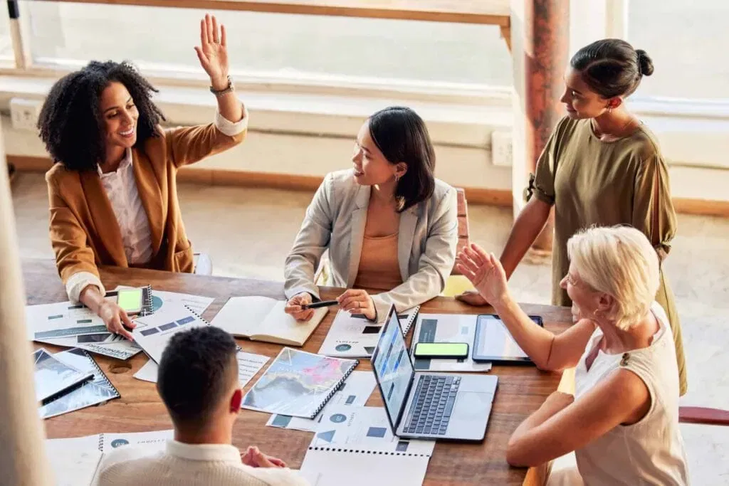 A diverse group of female professionals high-fiving and collaborating around a wooden table with laptops and documents.