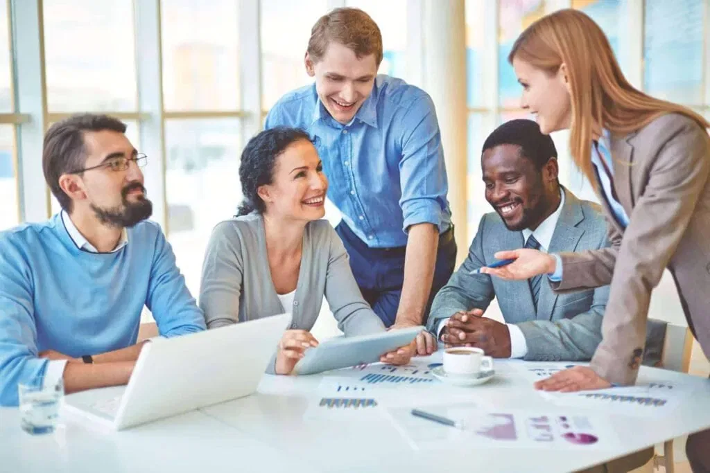 A diverse group of smiling professionals in a modern office setting discussing documents and a tablet during a strategy session.
