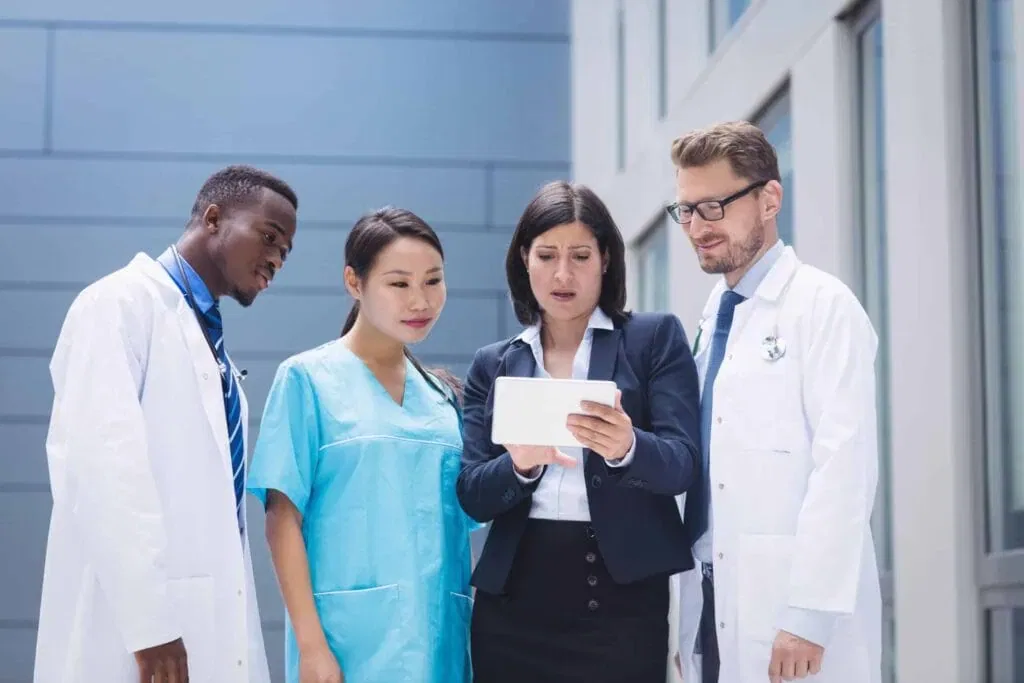 A diverse group of doctors and a nurse in lab coats and scrubs looking at a digital tablet held by a woman in a business suit.