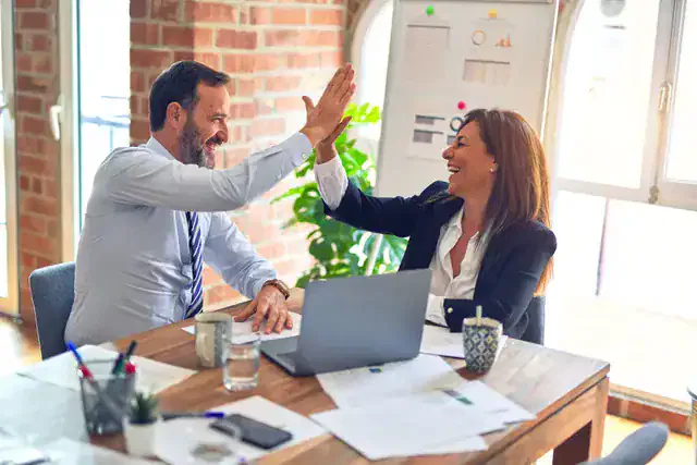 Two diverse hotel managers in professional attire giving a high-five and laughing during a productive meeting in a bright, modern Ocala office.
