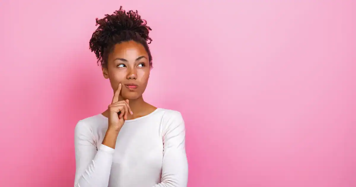 A young woman with curly hair tied up, looking thoughtful with her finger on her chin against a solid pink background with ample copy space.