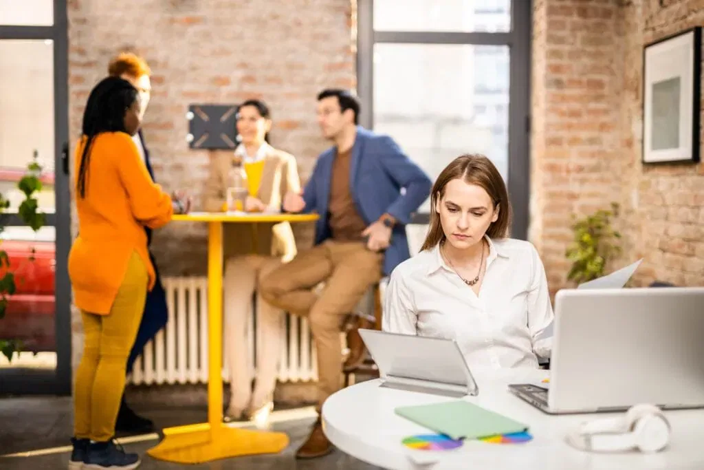 A focused female professional in a white shirt works at a round white table with a laptop and tablet. In the background of the brick-walled loft office, a diverse group of three colleagues stands around a high yellow table engaging in a collaborative discussion.