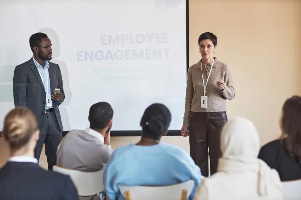 A professional, mature female HR manager standing and leading a corporate workshop. A male colleague stands beside a projection screen displaying "EMPLOYEE ENGAGEMENT: PERFORMANCE AND RESPONSIBILITIES" while a diverse group of employees listens in a bright conference room.