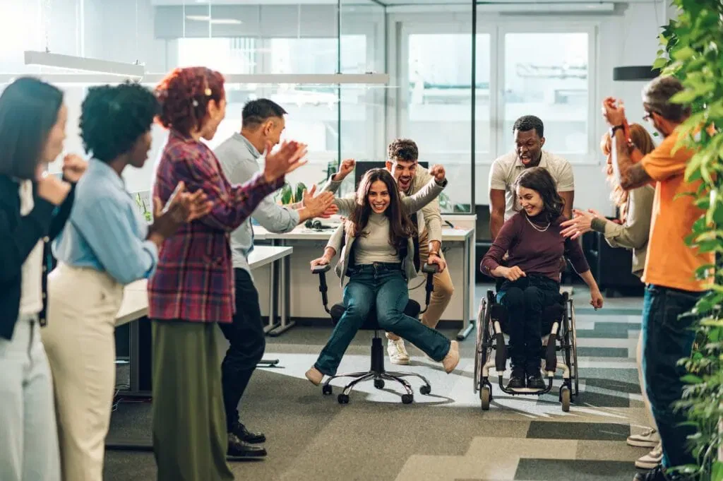 A diverse, multiracial business team cheering enthusiastically during a playful office chair race. One woman is being pushed in a rolling desk chair while another team member in a wheelchair participates, showcasing an inclusive, high-energy, and fun corporate culture.