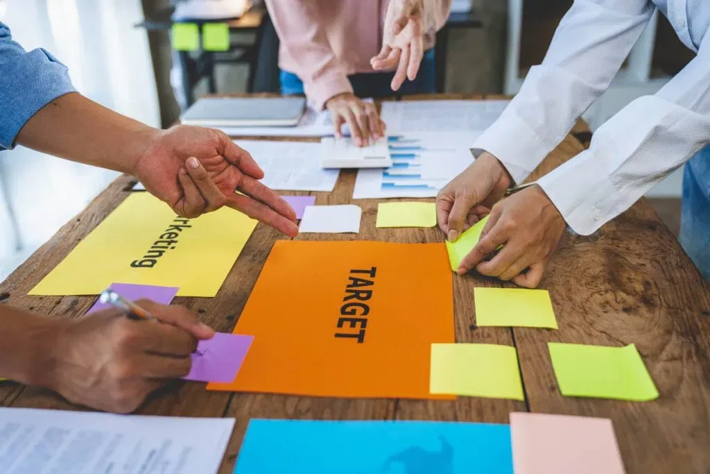 A close-up, high-angle view of business professionals' hands working around a wooden table with colorful sticky notes, a calculator, and papers labeled "Marketing" and "Target" during a strategy meeting.