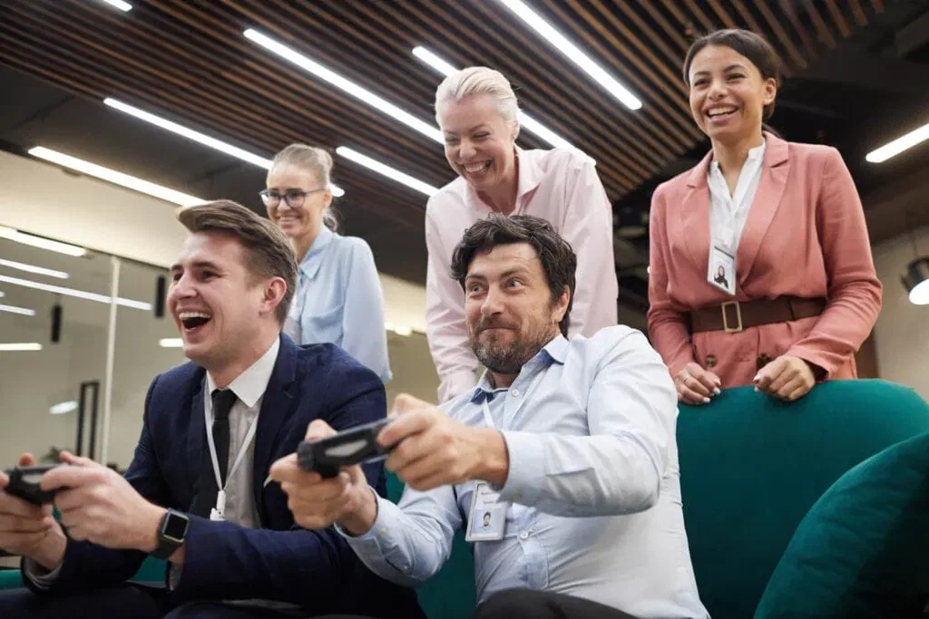 A diverse group of office professionals laughing and cheering while two men play video games with controllers in a modern corporate breakroom.