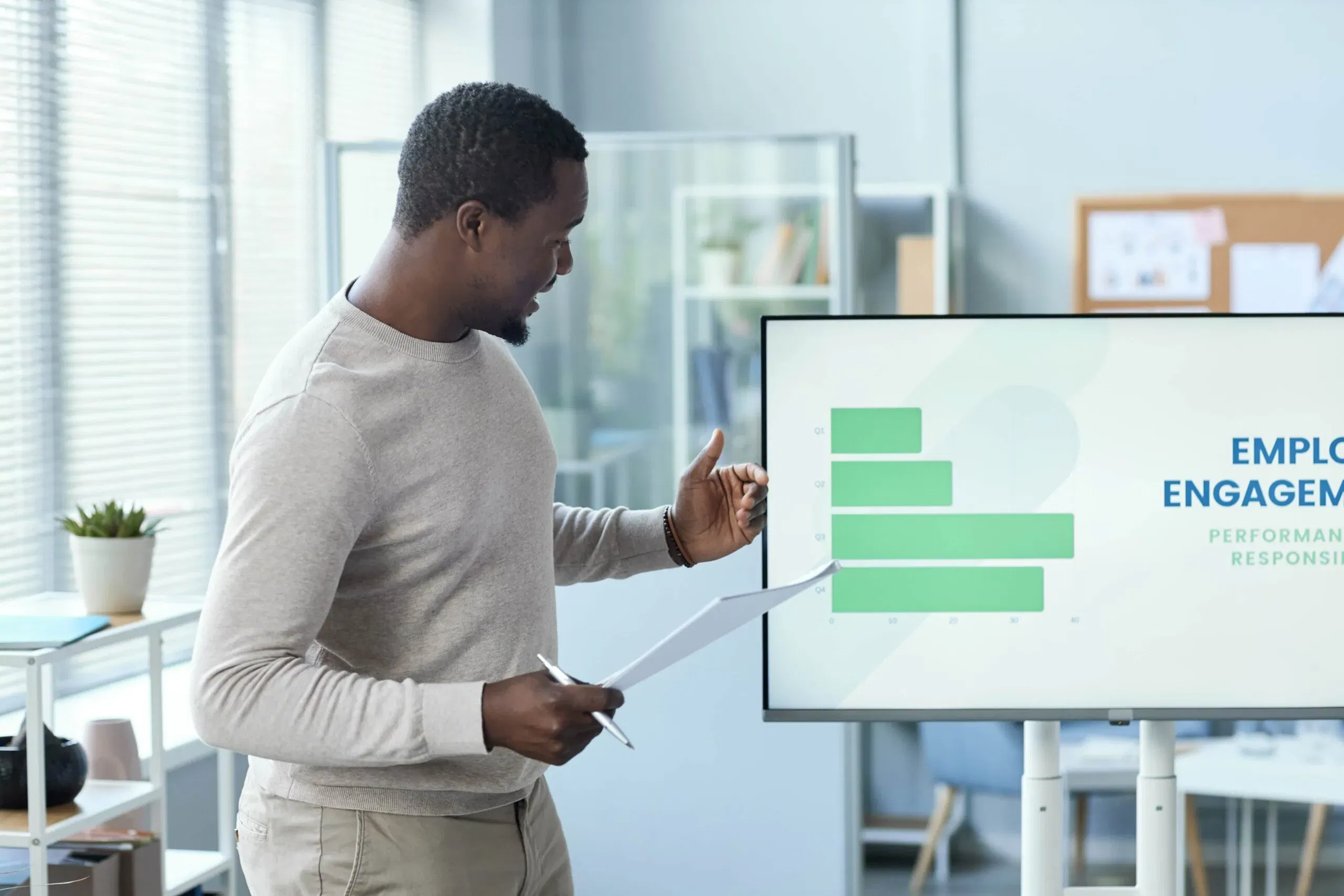 A professional Black man standing in a bright office, giving a presentation on employee engagement with a digital screen displaying a green bar chart titled Employee Engagement.