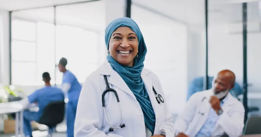 A smiling female doctor wearing a blue hijab and a white lab coat with a stethoscope around her neck, standing in a bright, modern hospital office with diverse colleagues in the background.