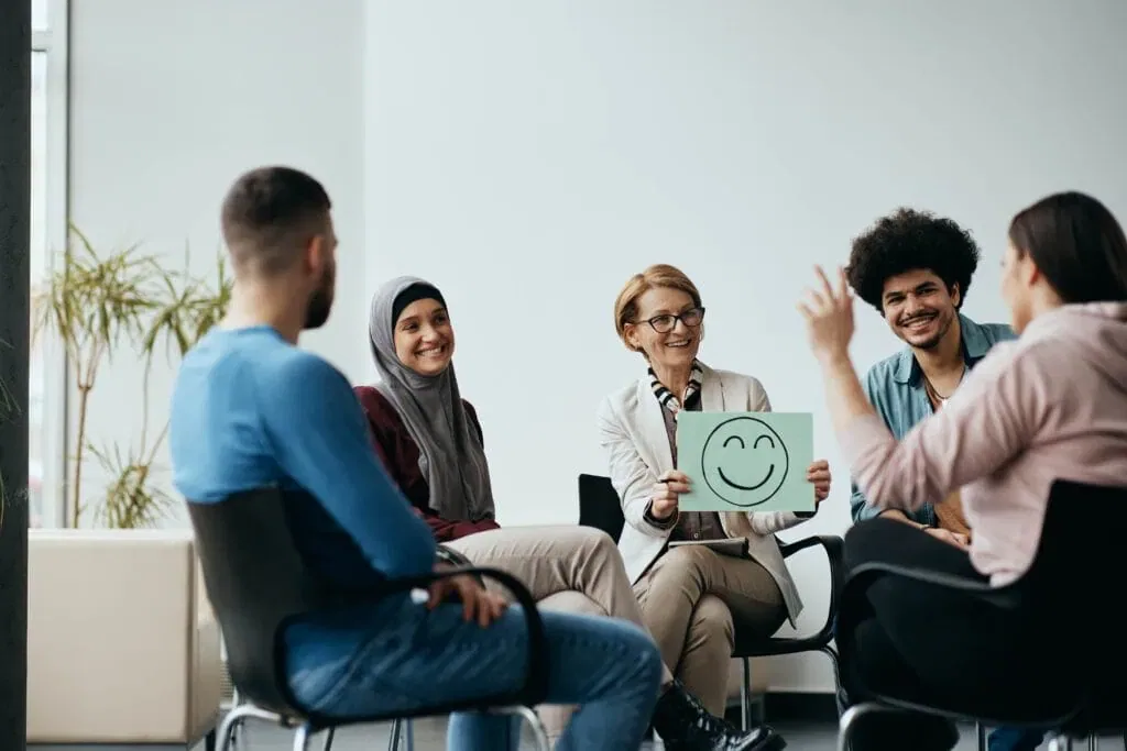 A smiling female therapist holding a green placard with a smiley face during a diverse group therapy session with young adults sitting in a circle.