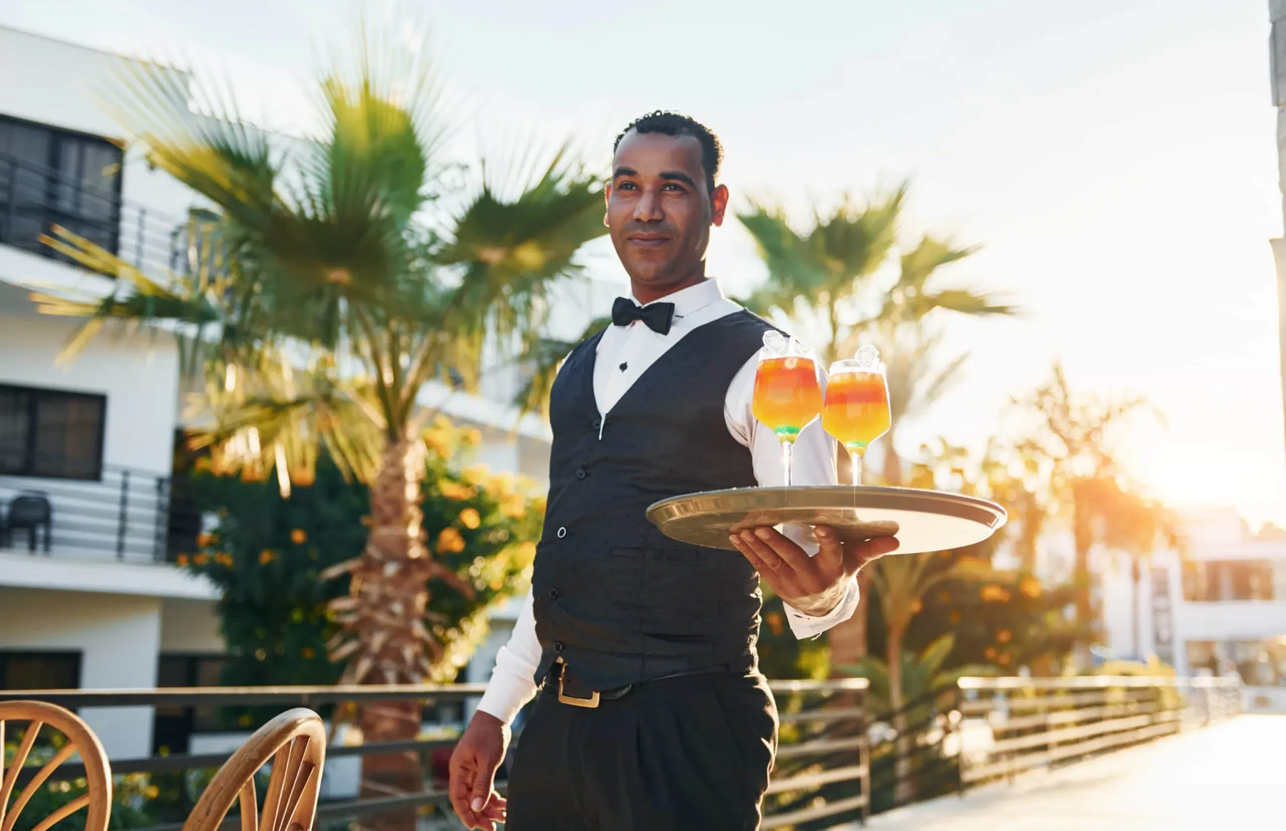 A professional hotel waiter in a formal vest and bowtie serving vibrant cocktails on a sun-drenched outdoor patio, providing elite guest service in Miami Beach, Florida.