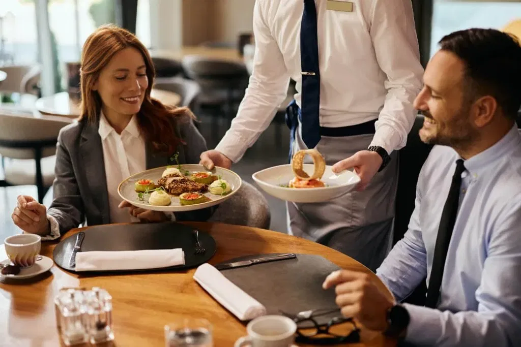 A professional waiter in a white shirt and blue tie serving gourmet plated meals to a businessman and businesswoman at a wooden table in an upscale restaurant.