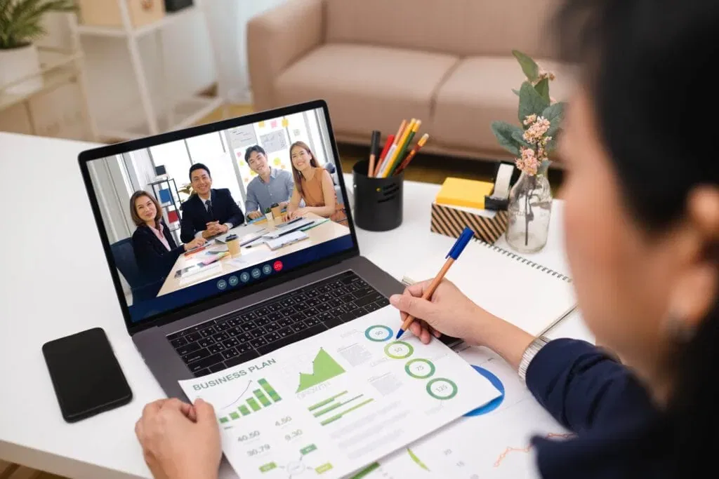 An over-the-shoulder view of an Asian woman working from home, presenting a business plan with green charts during a video conference call with colleagues on a laptop.